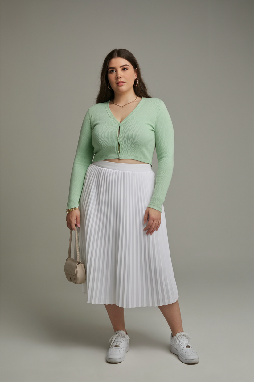 Plus-size woman in a pastel green cardigan with a white pleated midi skirt and white sneakers, standing against a gray background.