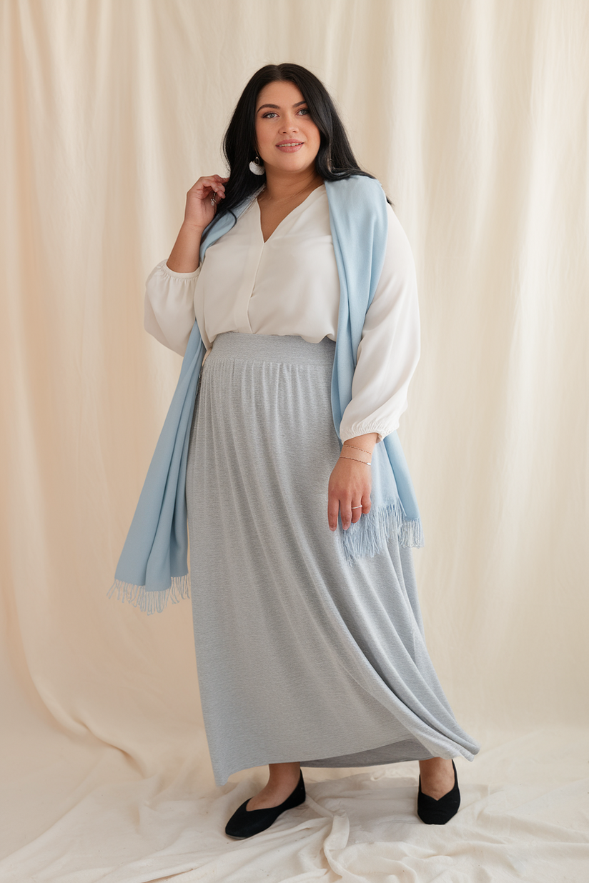 Plus-size woman in a gray maxi skirt and white blouse with a blue scarf, standing against a cream-colored background.