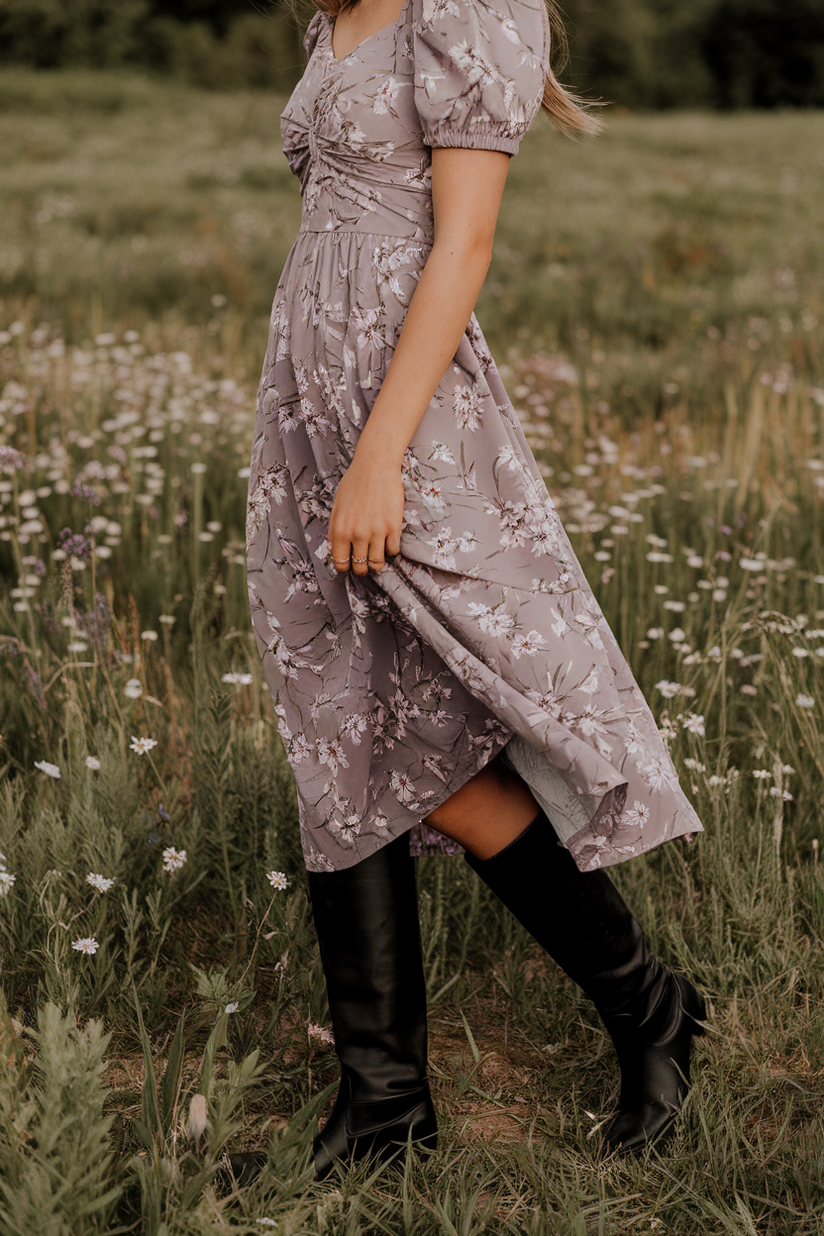 Woman in lavender floral dress and black knee-high boots in a wildflower field.