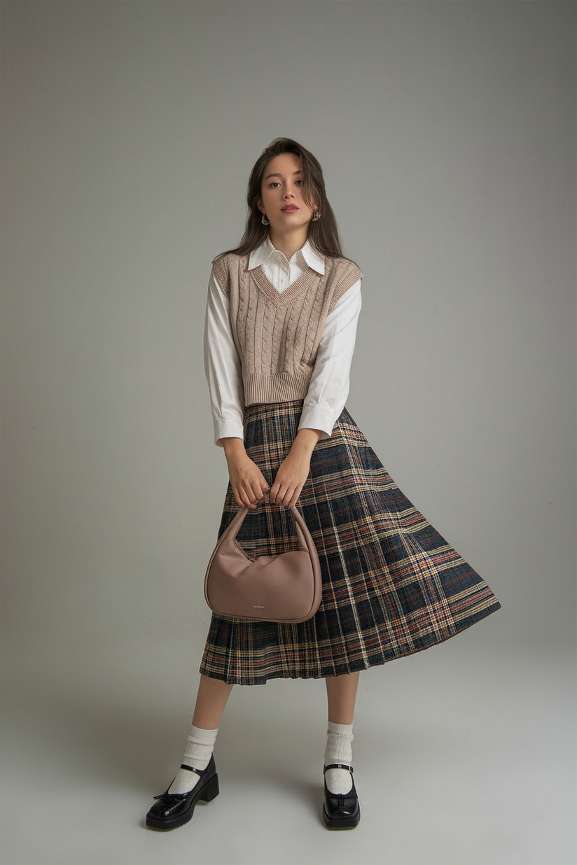 A woman wears a plaid pleated skirt, a knit vest, and black Mary Janes, embodying a classic preppy aesthetic.